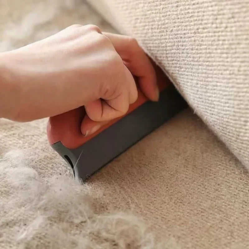 A hand using a terracotta and grey silicone pet hair remover tool to clear fluff from a beige fabric sofa.