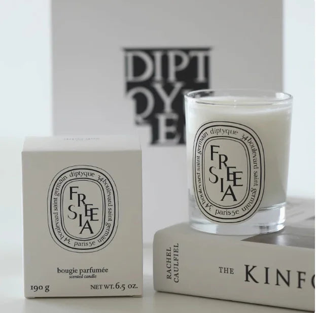 Freesia scented candle in a clear glass jar with a white box, featuring black oval branding on a cream background.