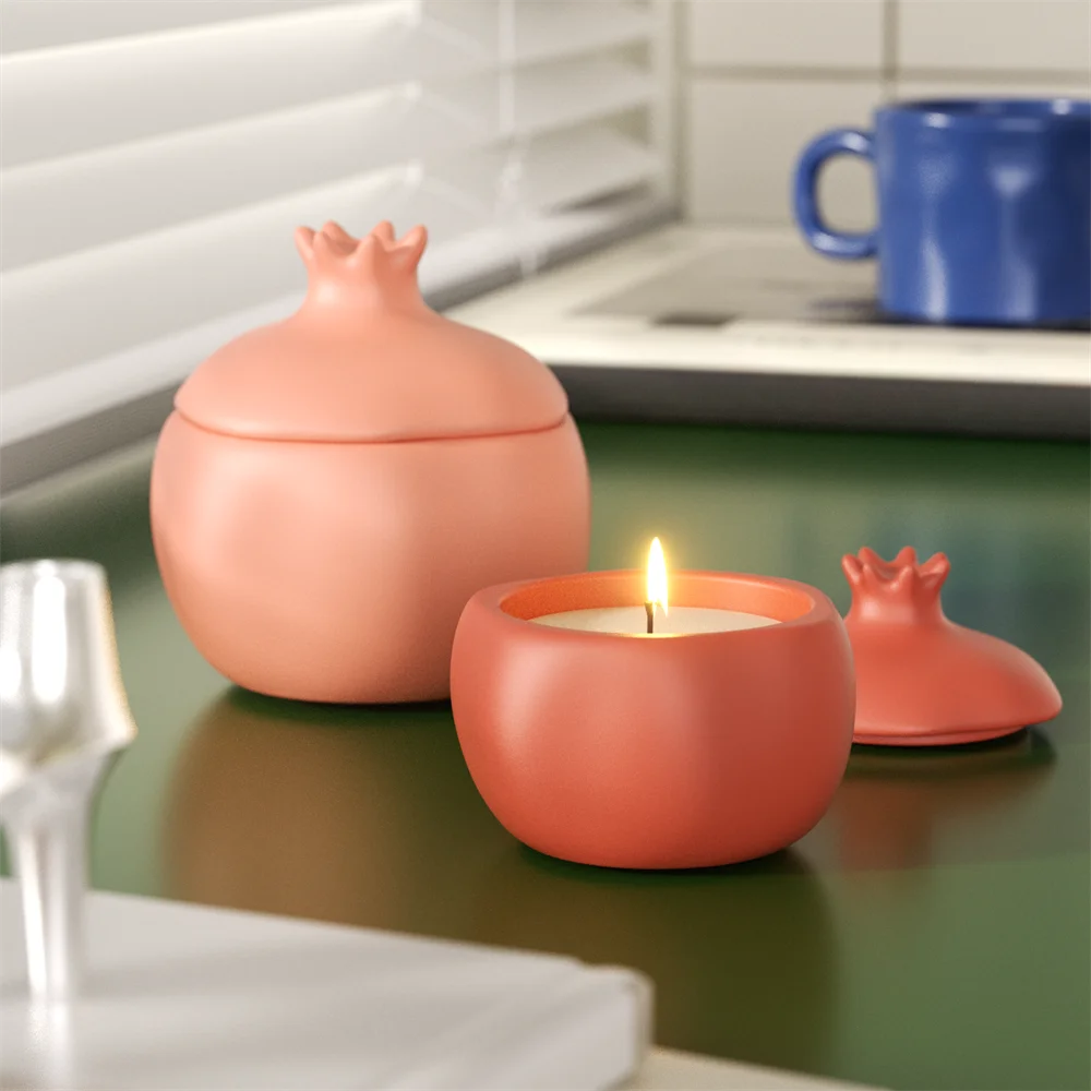 Pink and terracotta pomegranate-shaped candle jars with lids, one burning.