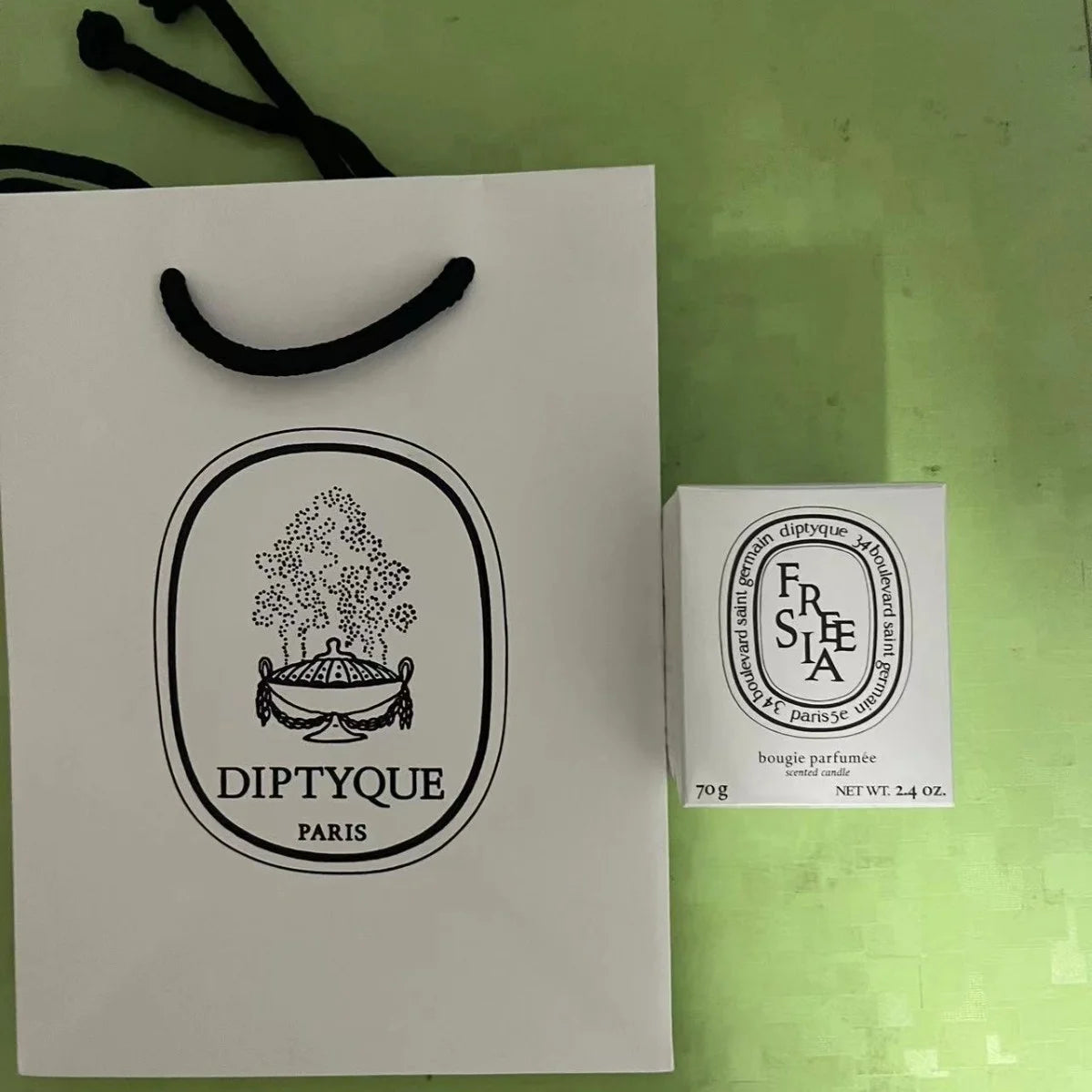 White branded shopping bag with black handles next to a 70g white box containing a luxury scented candle.