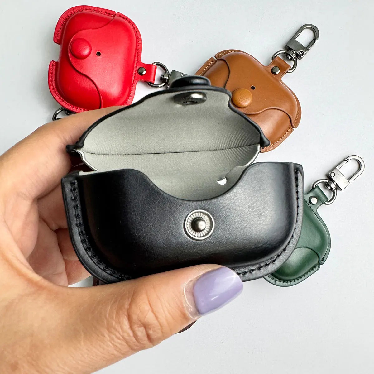 Leather AirPods Pro cases in black, red, tan, and green, featuring snap-button closures and metal keychains.