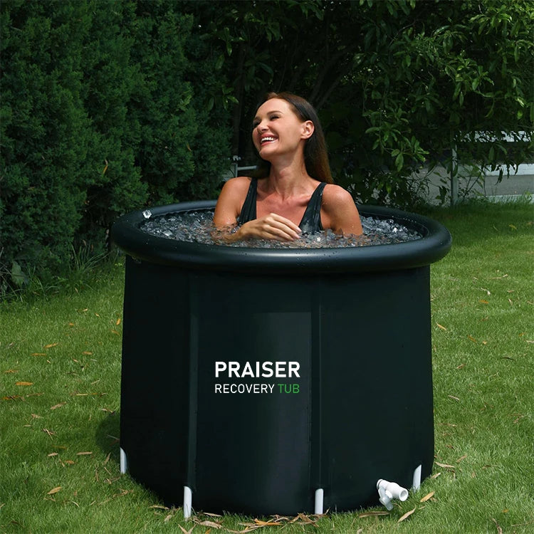A smiling woman in a black swimsuit sits in a black round PRAISER RECOVERY TUB filled with ice water on a lawn.