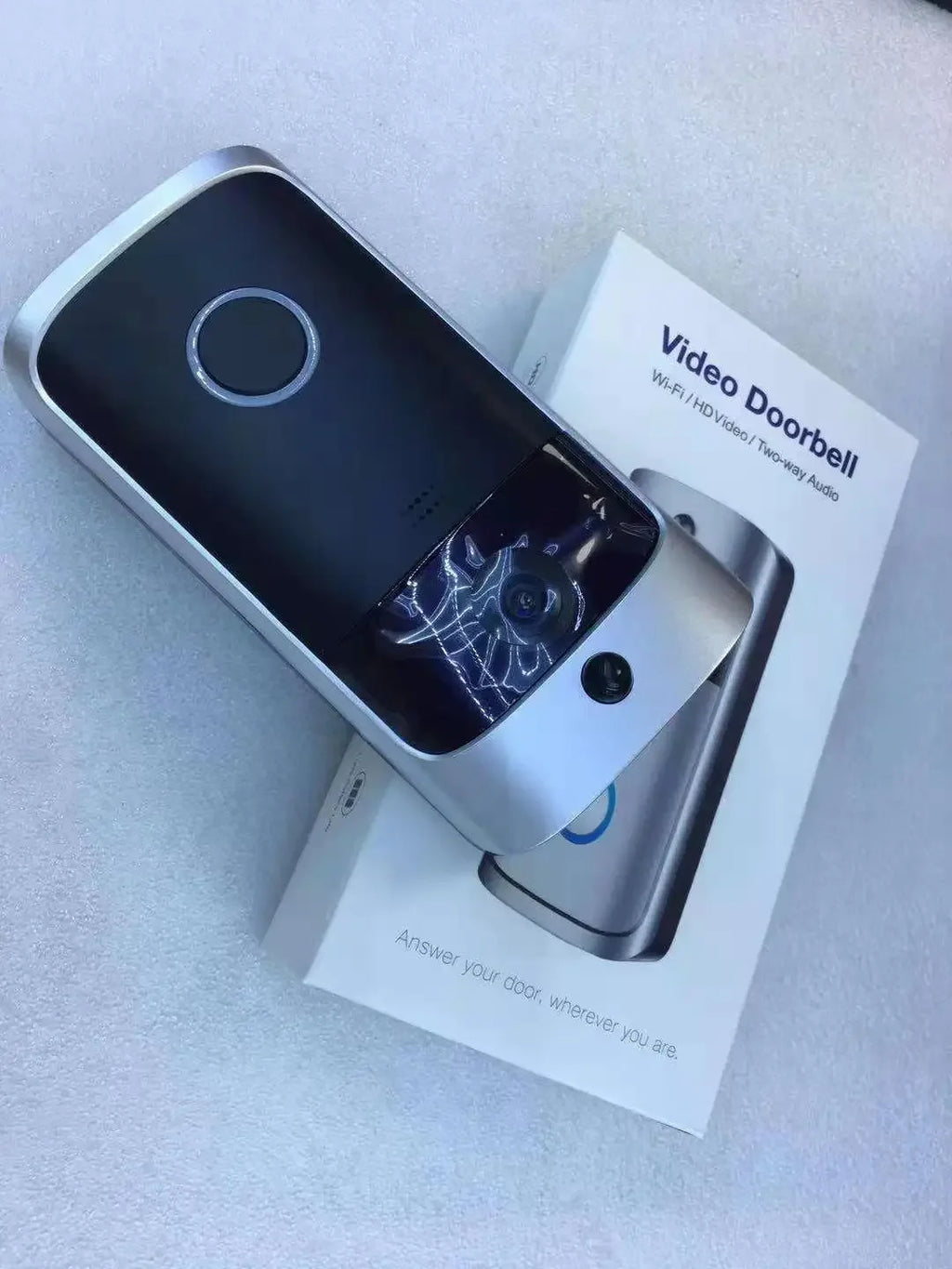 Silver and black smart wireless video doorbell with camera lens, mounted on its white retail packaging.
