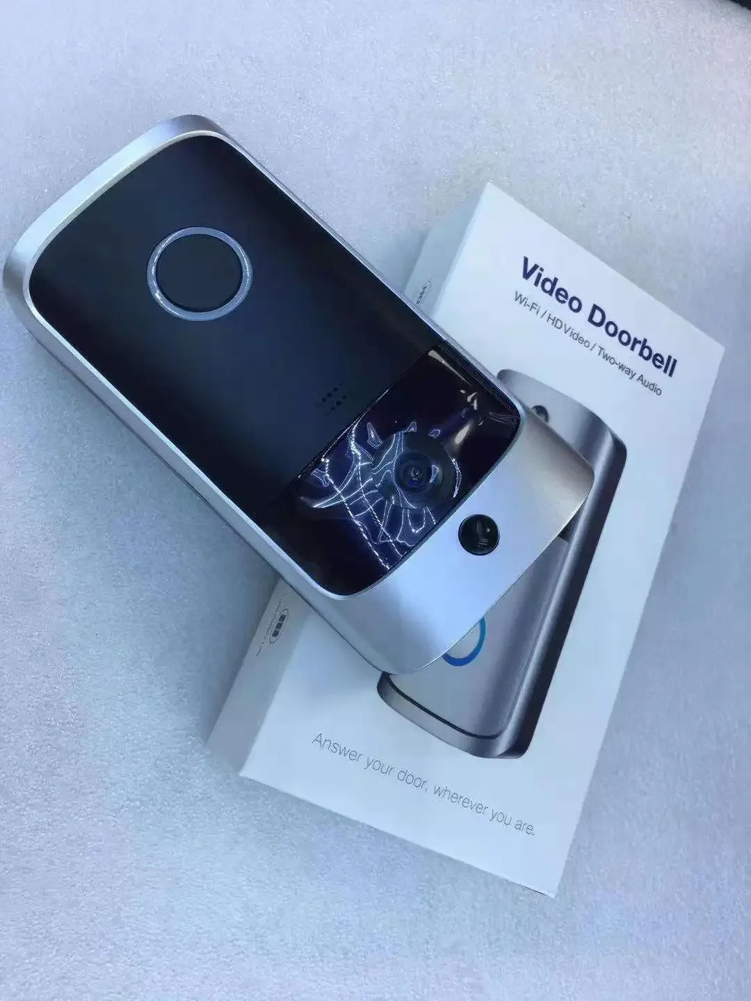 Silver and black smart wireless video doorbell with camera lens, mounted on its white retail packaging.