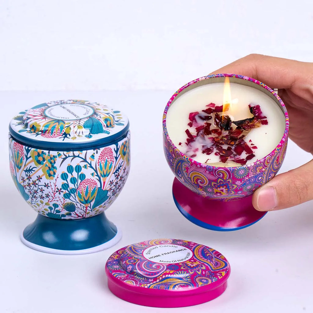 Two decorative metal-tin candles with colourful floral and paisley patterns, one lit with botanical wax toppings.
