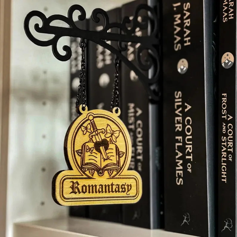A wooden, hanging sign labelled ‘Romantasy’ with a sword, book, and butterfly design, suspended from a black bracket.
