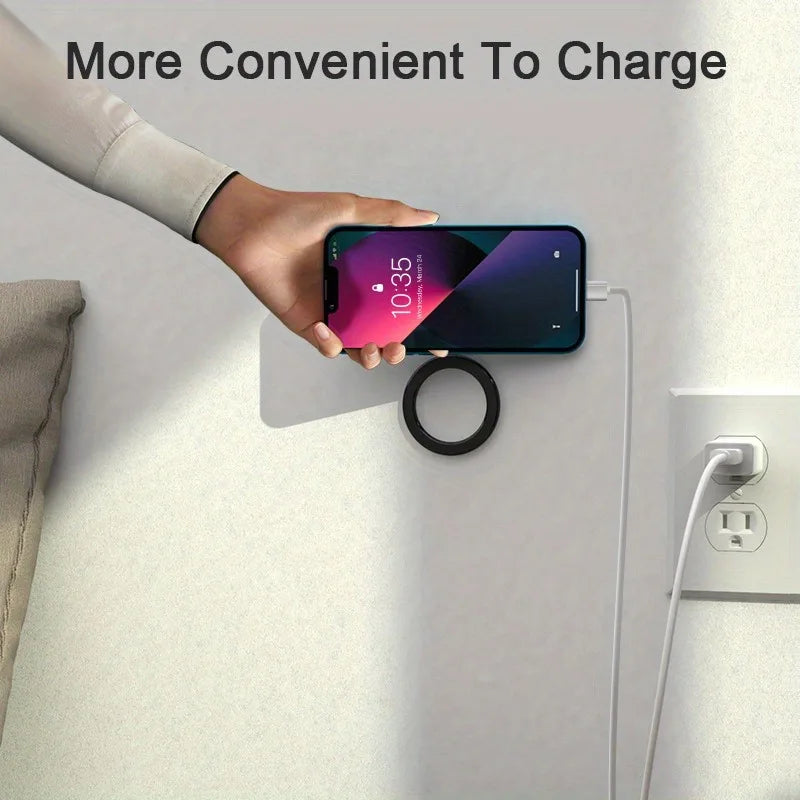 A hand holding a smartphone against a wall-mounted, round, black magnetic wireless charger plugged into an outlet.