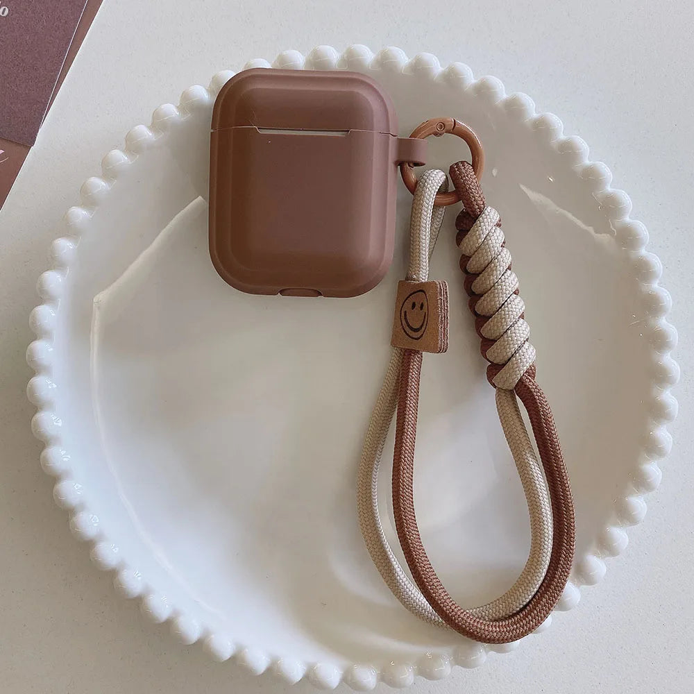 Brown matte silicone case for AirPods with a two-tone knotted rope lanyard featuring a smiley face tag.