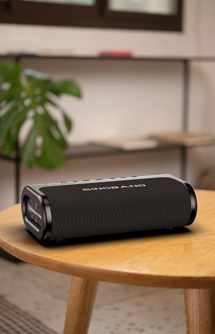 Portable black rectangular Bluetooth speaker with a mesh fabric finish, sitting on a wooden side table.