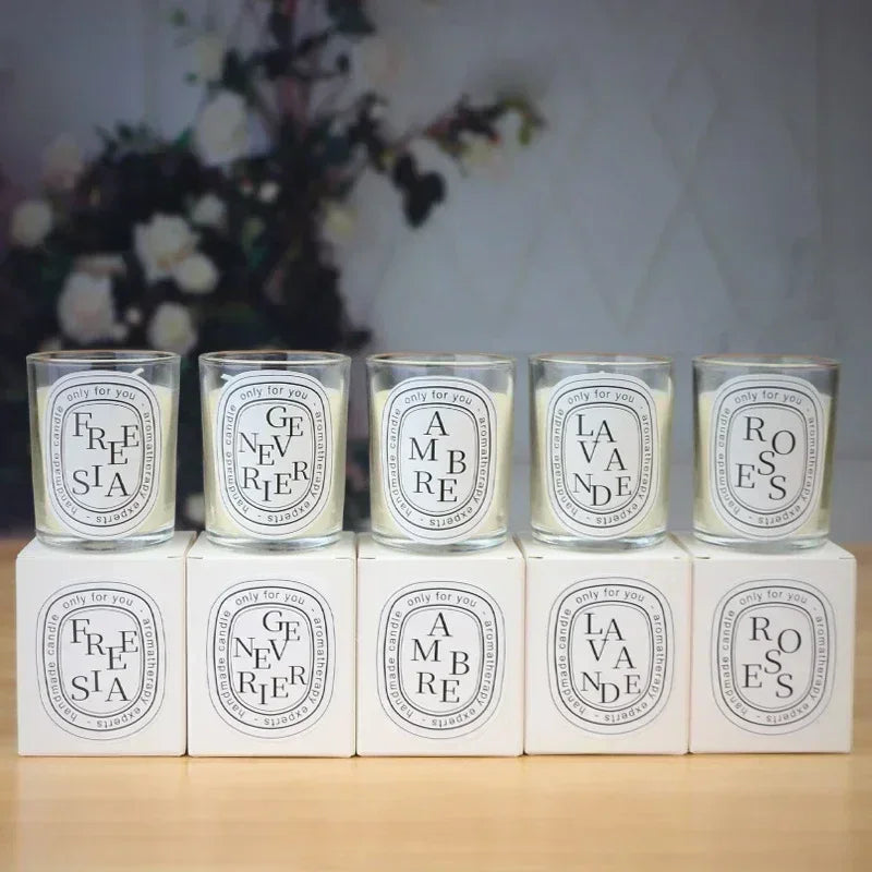 Five scented candles in glass jars with white labels, arranged on matching white gift boxes.