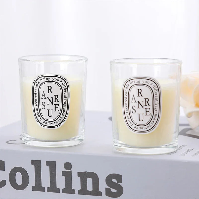 Two scented candles in clear glass tumblers with oval labels on a book, featuring cream wax and a minimalist design.