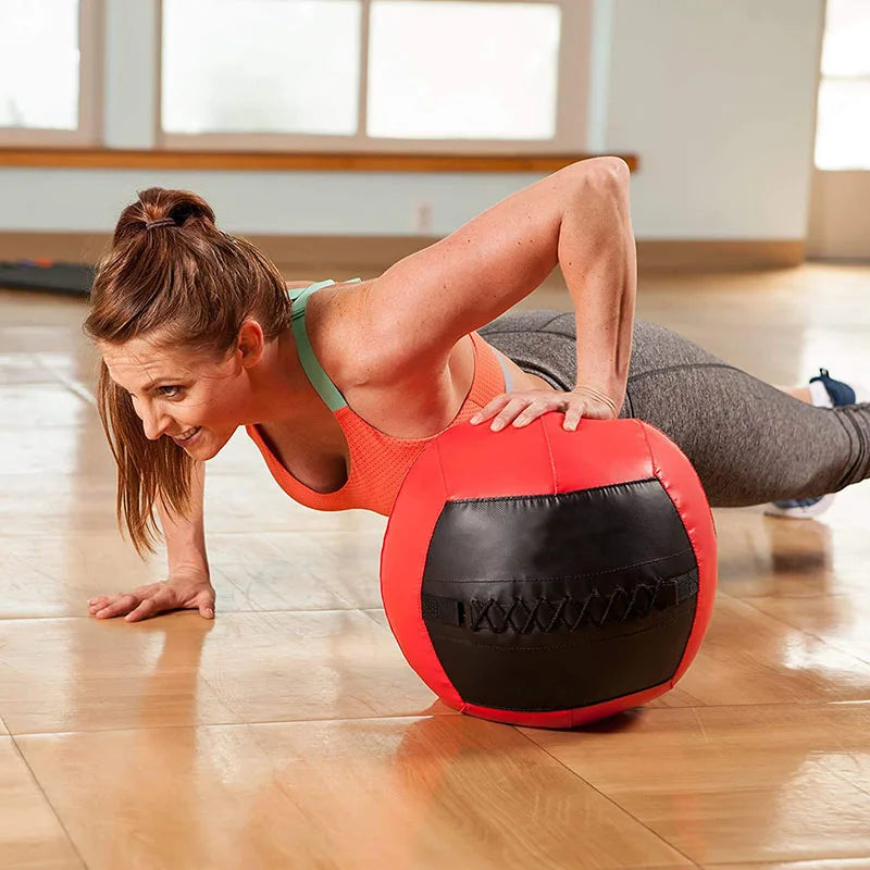 A woman does a push-up with one hand on a red and black medicine ball on a wooden floor.