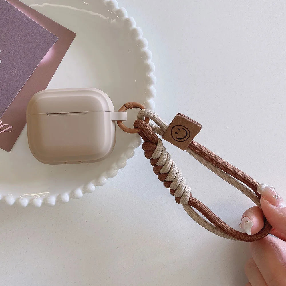 Beige silicone AirPods case with a braided two-tone lanyard and smiley face charm.