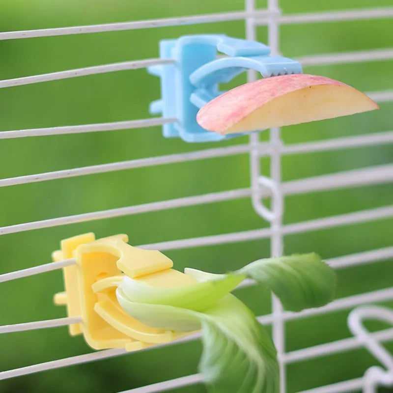 Blue and yellow plastic bird cage clips holding apple slice and leafy greens on white wire bars.