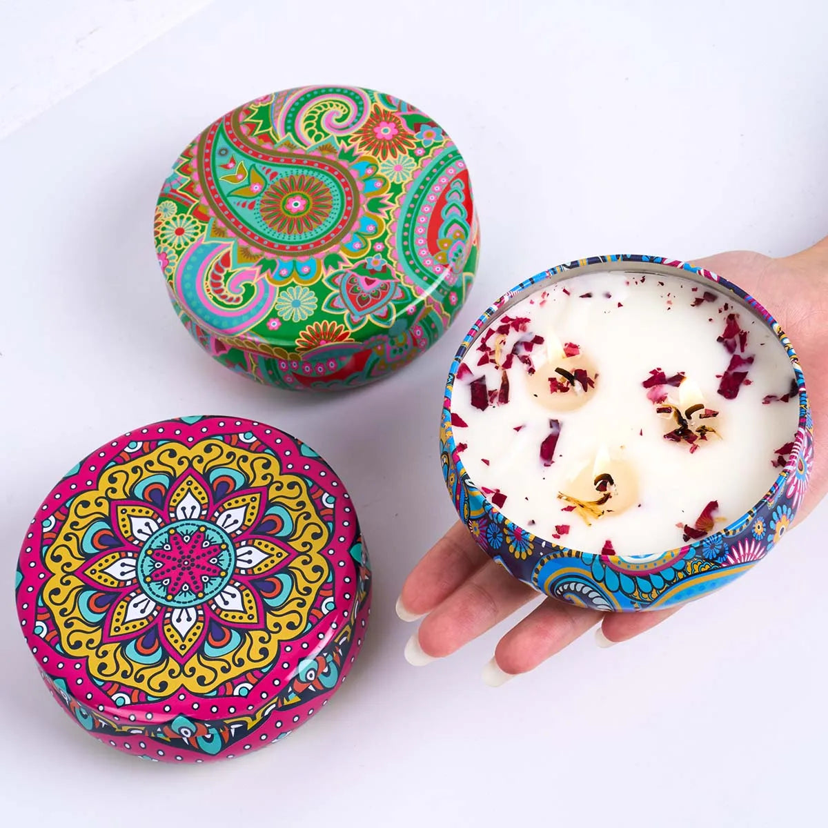 Three round, colourful tins with intricate floral and paisley patterns; one open candle features wax and dried petals.