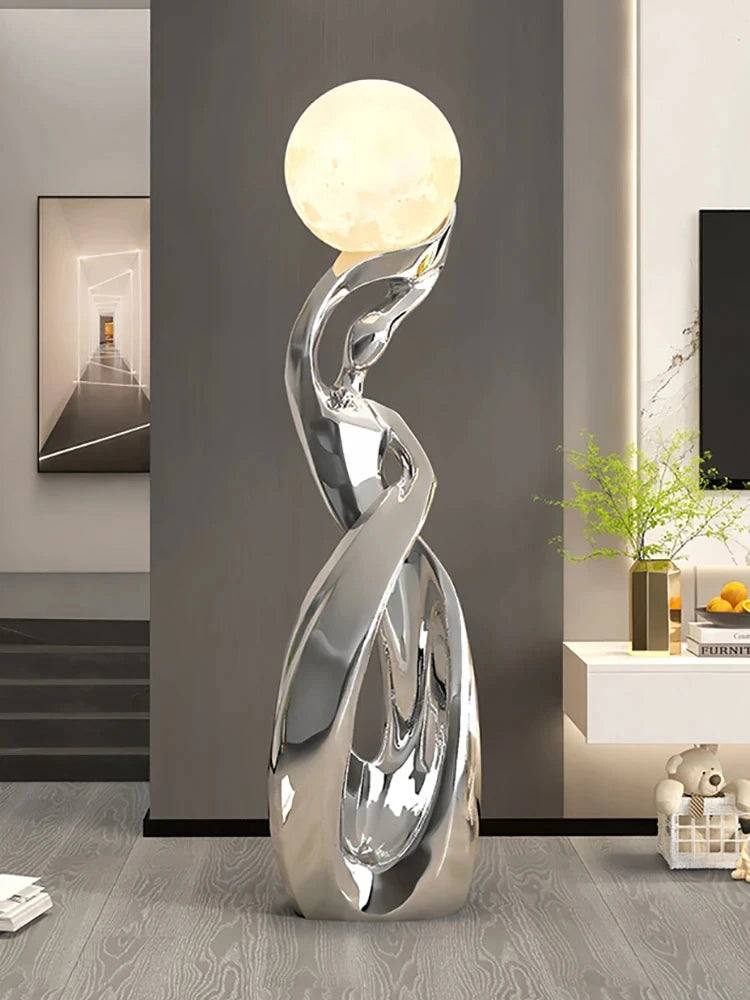 Modern abstract floor statue in polished chrome with a glowing, moon-like orb lamp at the top.