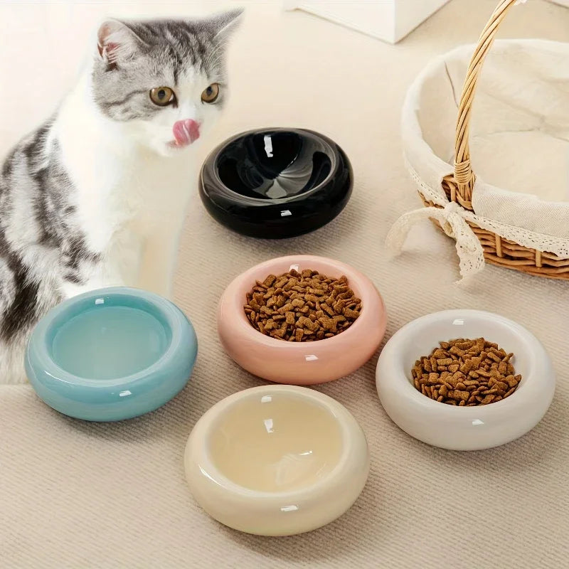 A grey and white cat beside five smooth, round ceramic feeding bowls in black, blue, pink, white, and cream.
