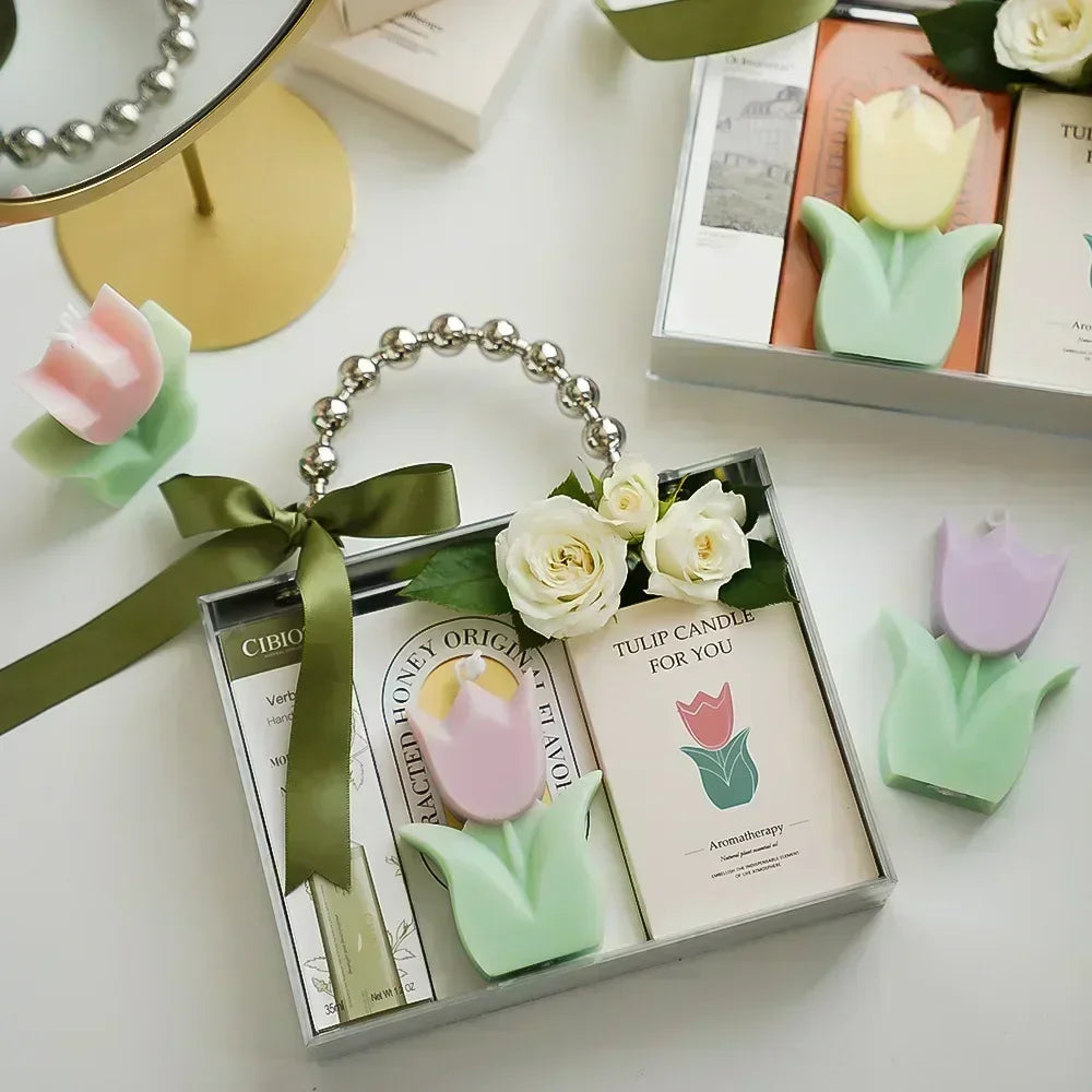 Gift set featuring pastel-coloured tulip candles, a decorative green ribbon, and white roses in a clear carry box.