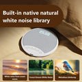 White, round Bluetooth pillow speaker with a grey fabric top, digital clock, and control buttons on a bedding surface.