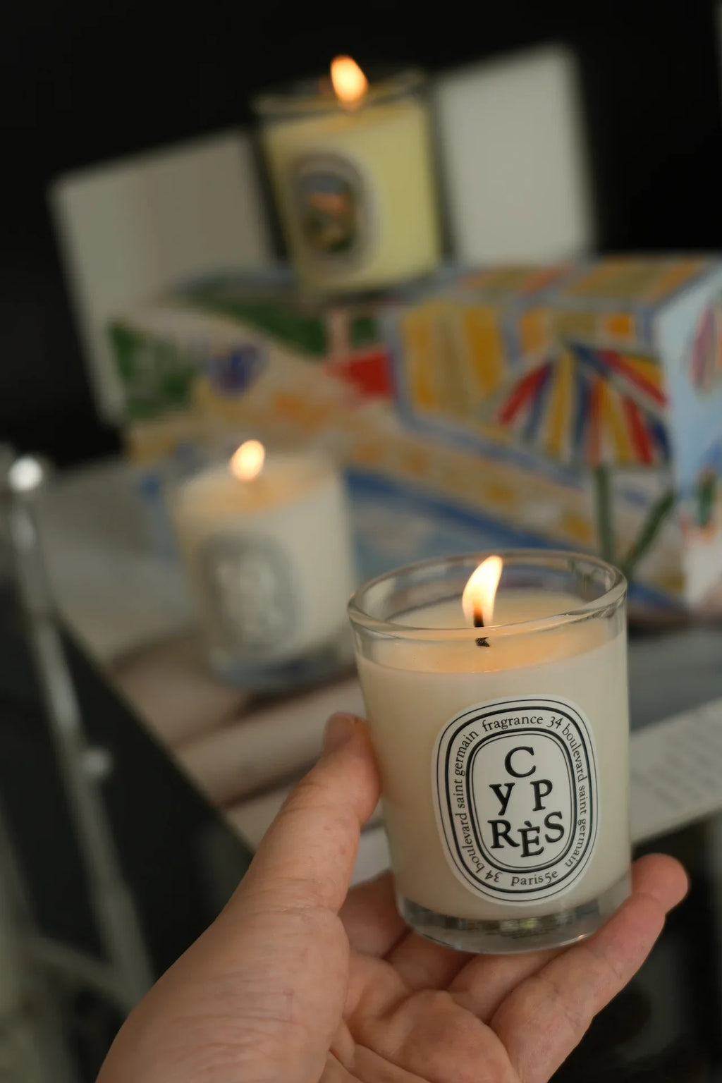 Hand holding a burning white wax candle in a clear glass jar with a decorative oval label, with blurred candles behind.