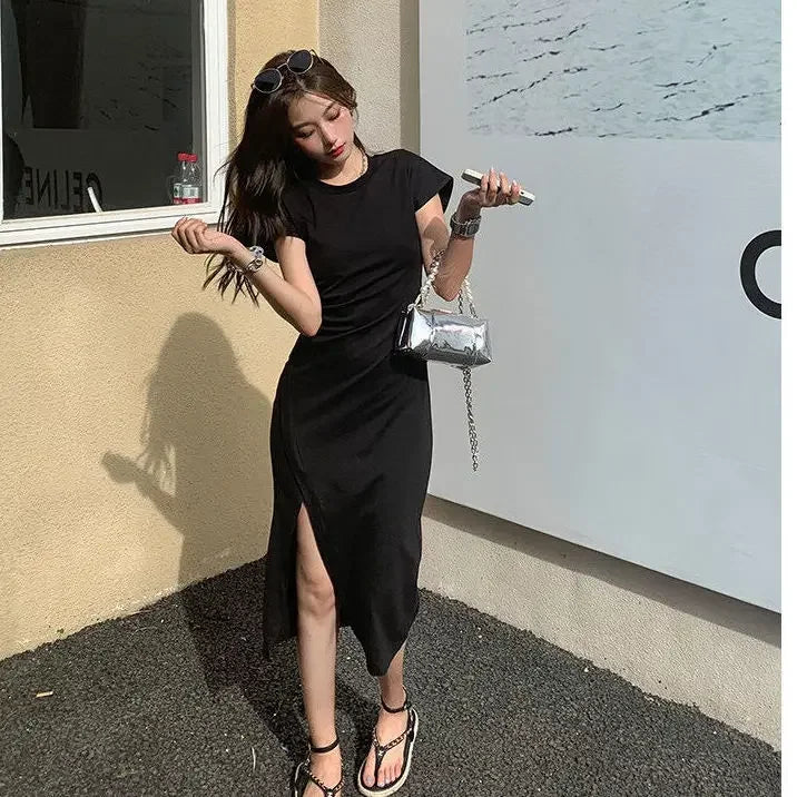 A woman wearing a black short-sleeved midi dress with a side slit, paired with a metallic silver handbag.