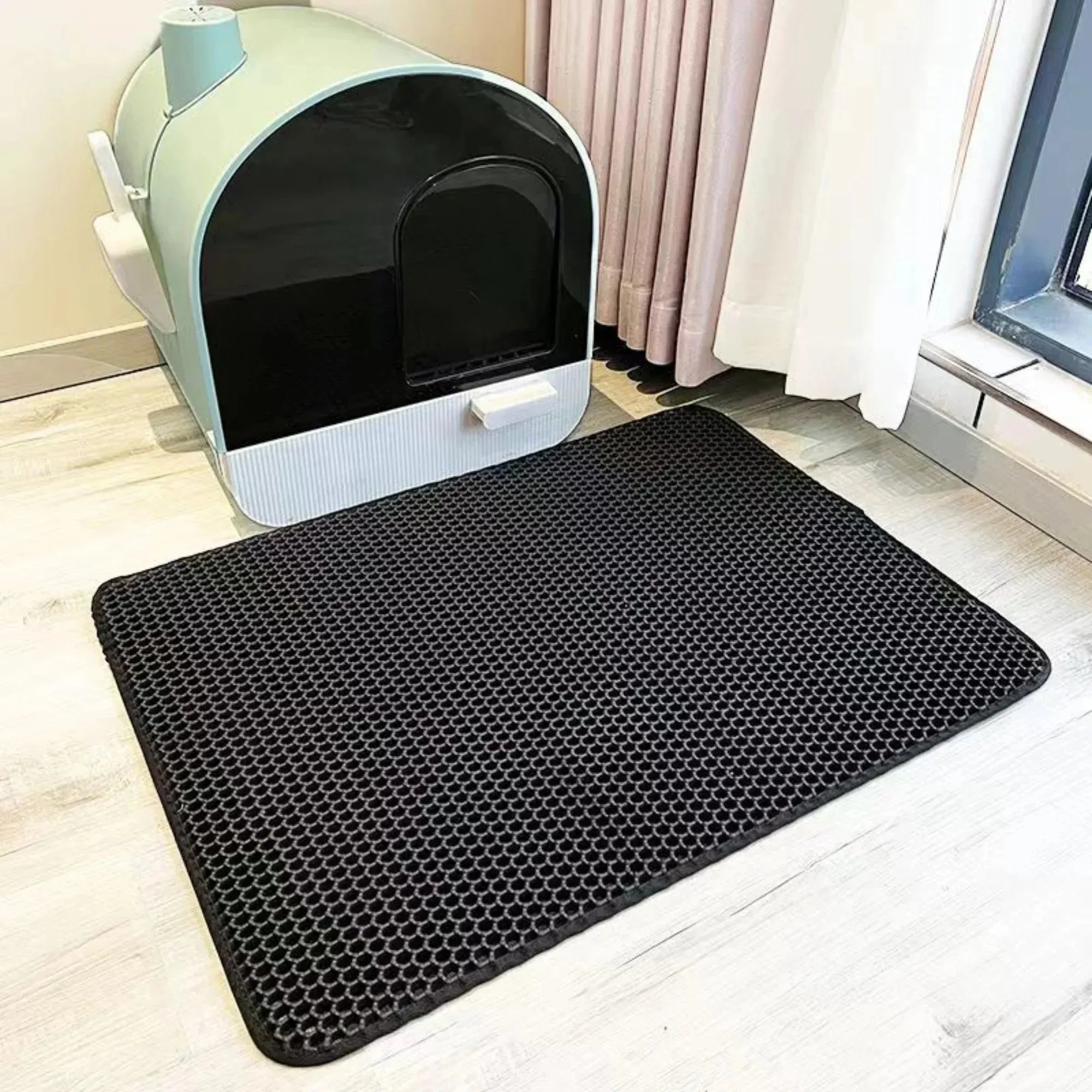 Black waterproof cat litter mat with a honeycomb texture design, placed on a light wood floor by a litter tray.