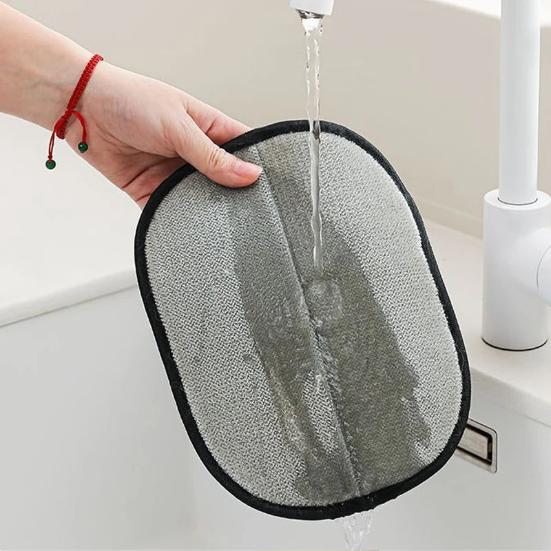 A grey, oval-shaped cleaning sponge with a black trim being held and rinsed under a kitchen tap.