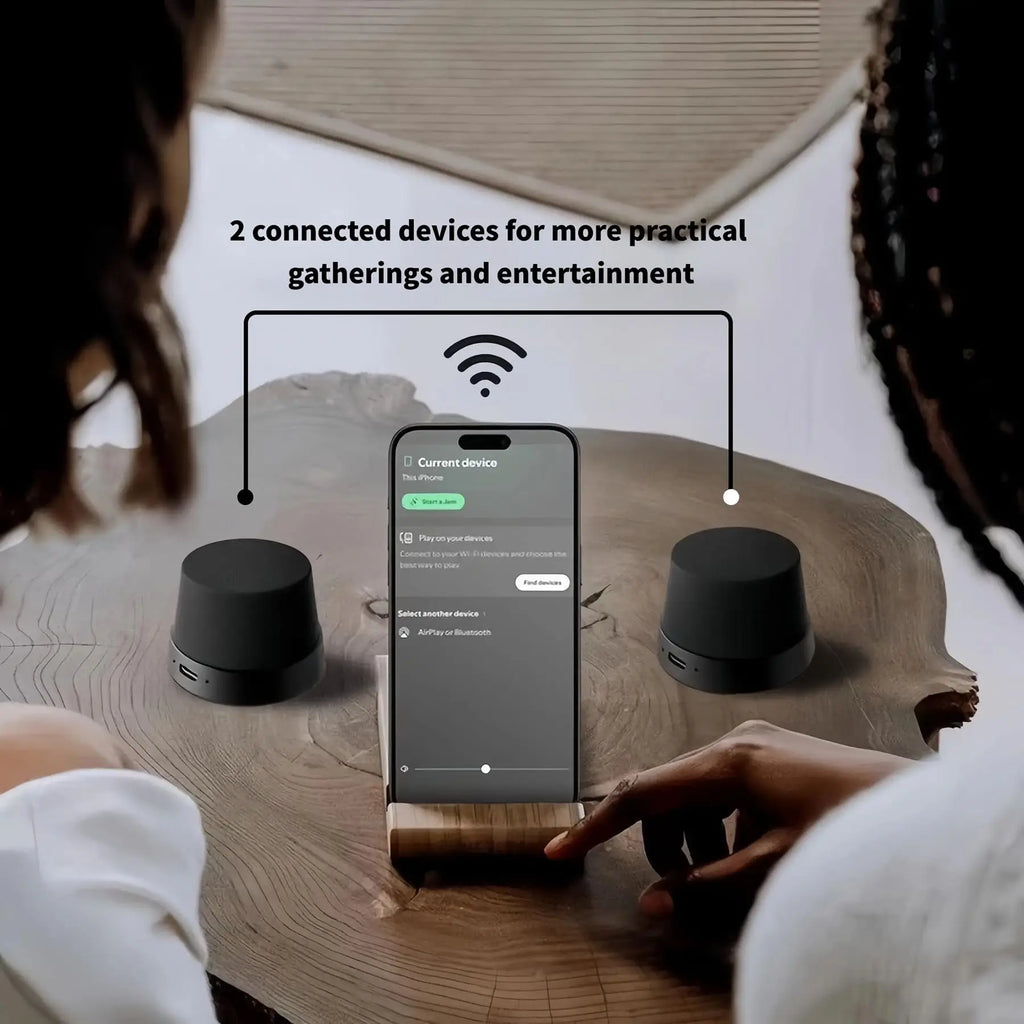 Two sleek, matte black wireless portable speakers shown connecting to a smartphone for dual-audio playback.