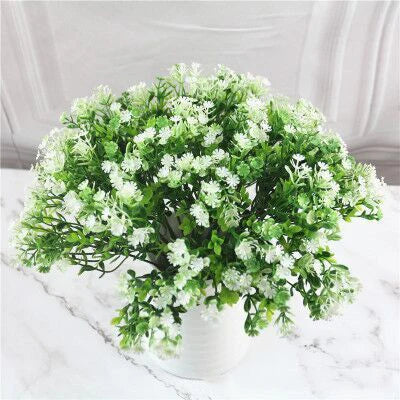 A lush bouquet of artificial green leaves and small white flowers in a white pot on a marble-patterned surface.