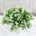 A lush bouquet of artificial green leaves and small white flowers in a white pot on a marble-patterned surface.