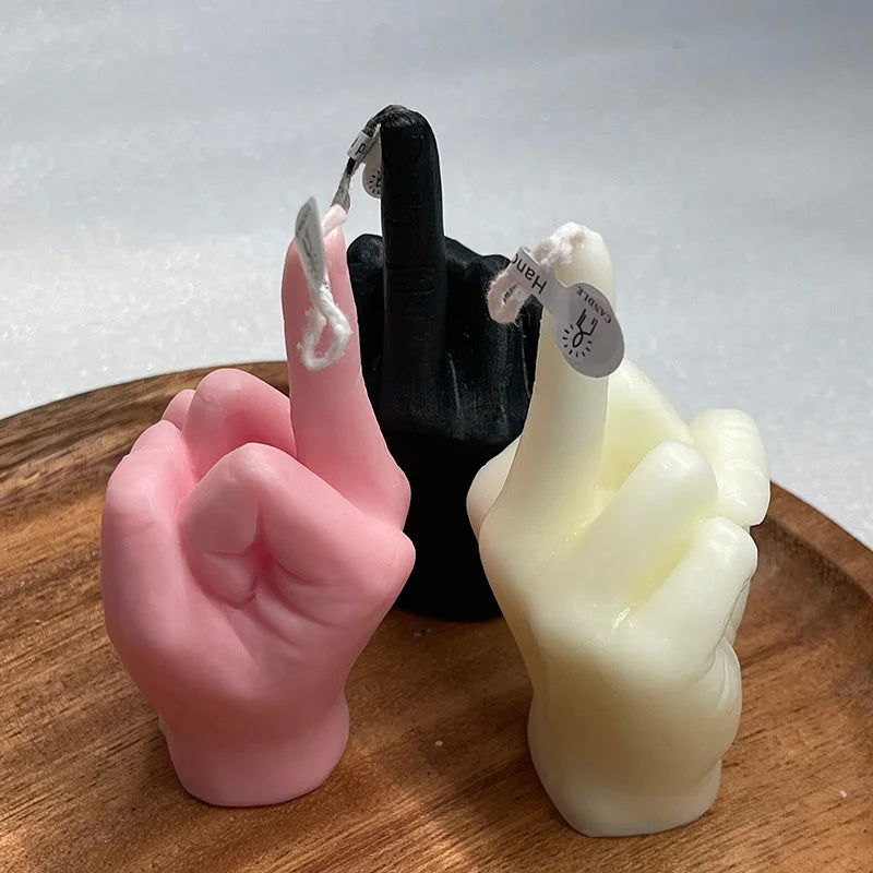 Three hand-shaped candles in pink, black, and white on a wooden board, each with a wick on the middle finger.