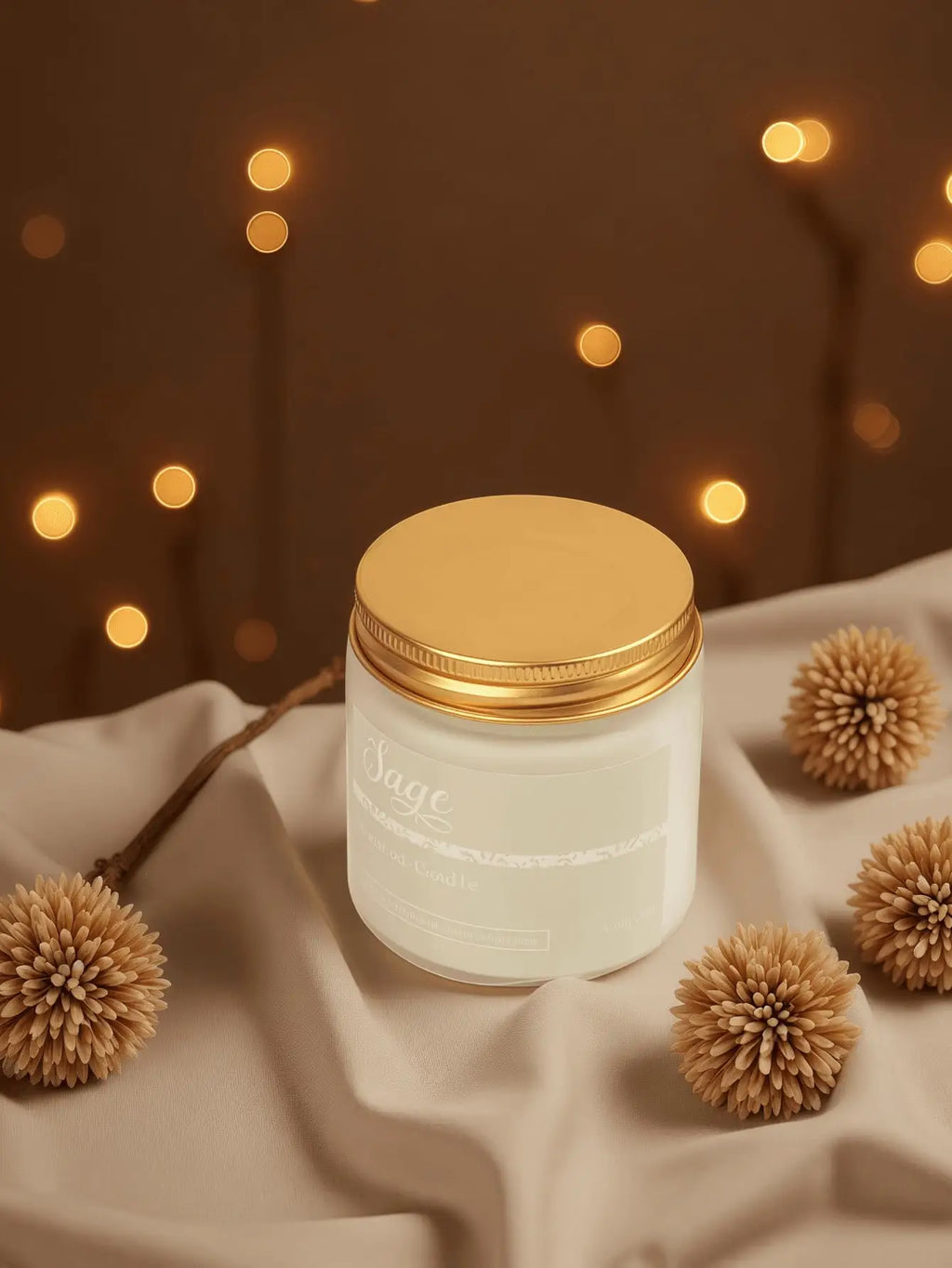 Sage-scented candle in a white glass jar with a metallic gold lid, set against warm fairy lights and fabric.