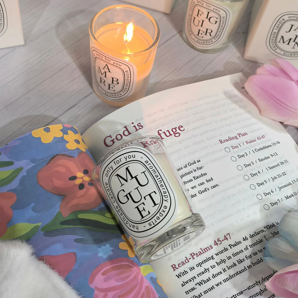 A lit glass candle with a classic oval label resting on a floral patterned book page, surrounded by soft-focus blooms.