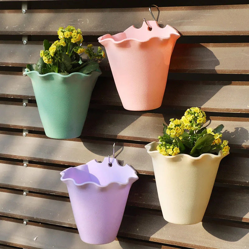 Four pastel, ruffle-rimmed plastic wall planters hang on a brown slat wall. Two contain small yellow flowering plants.