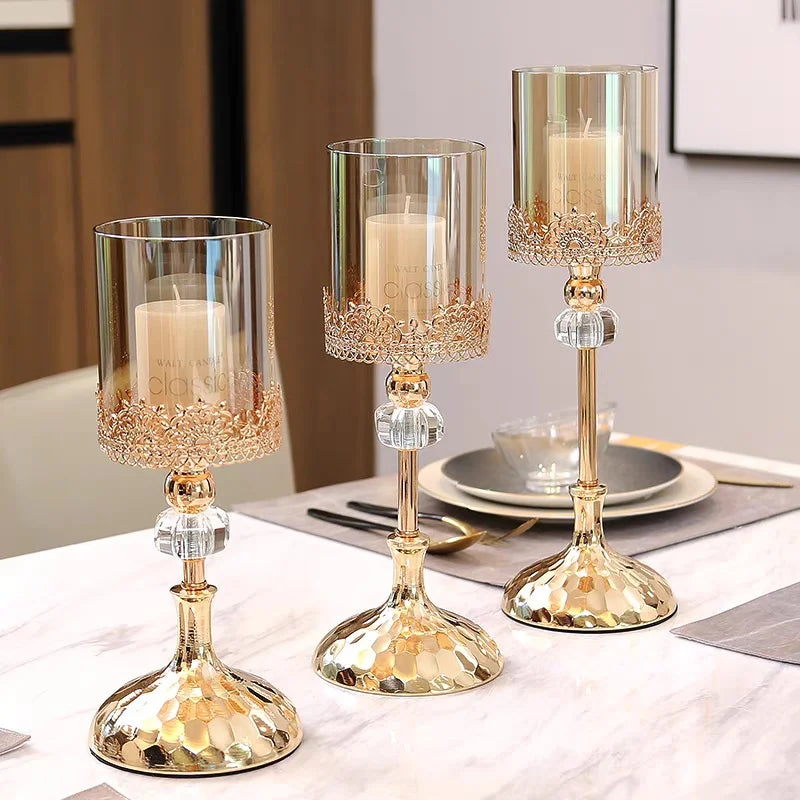 Three gold and crystal candle holders with glass cylinders and white pillar candles on a table.
