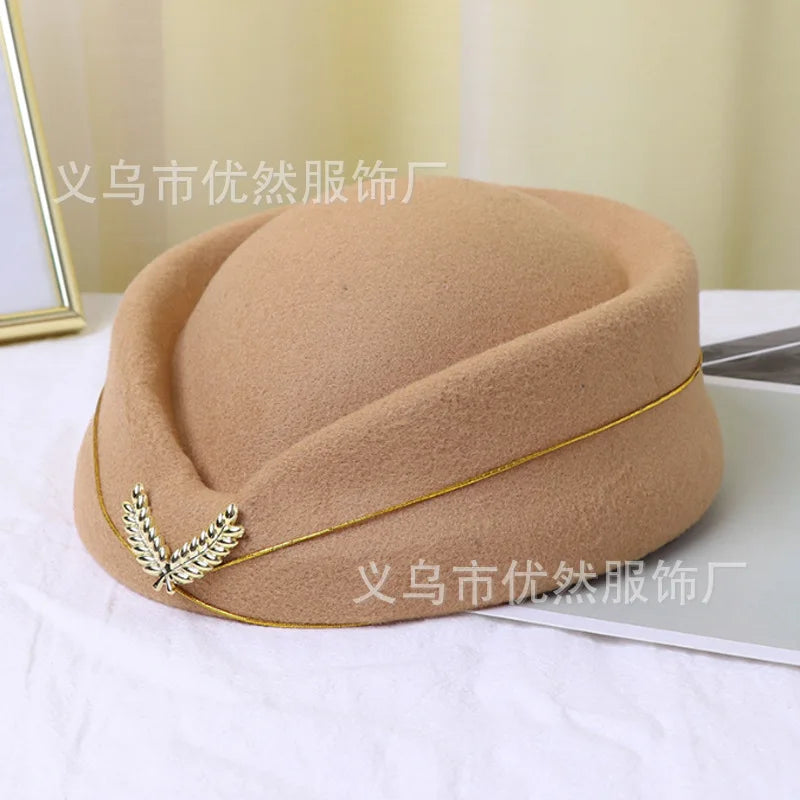A beige felt vintage-style stewardess pillbox hat, finished with a gold cord trim and decorative leaf emblem.