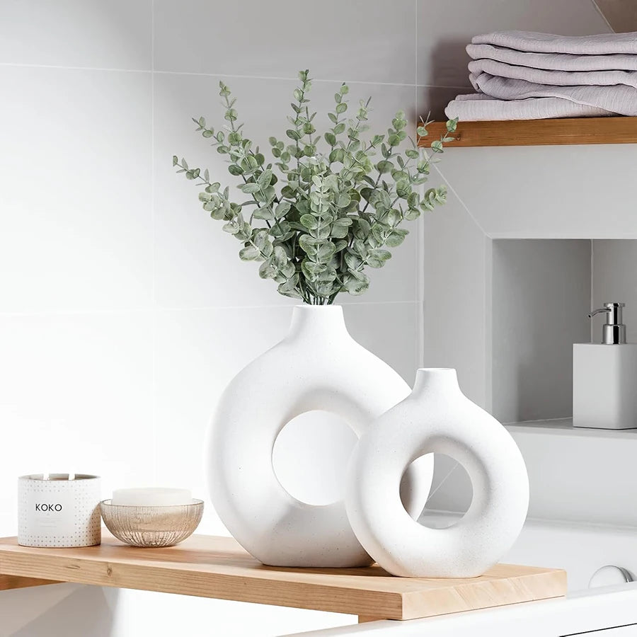 Two white, modern circular plastic vases with a matte finish, displayed on a wooden shelf with eucalyptus stems.