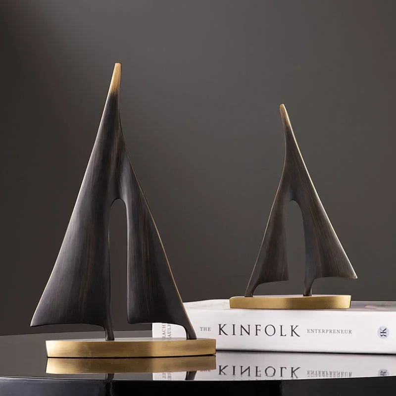 Two abstract, dark bronze-finish sailboat ornaments on gold-toned bases, styled with minimalist, modern lines.