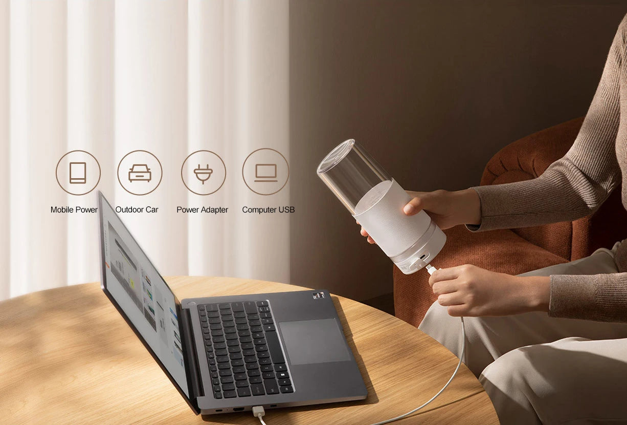 A person charges a minimalist white, portable, cylindrical USB blender next to a laptop on a wooden table.