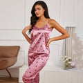 Woman wearing a dusky pink satin pyjama set with a black heart print, featuring a camisole top and matching trousers.