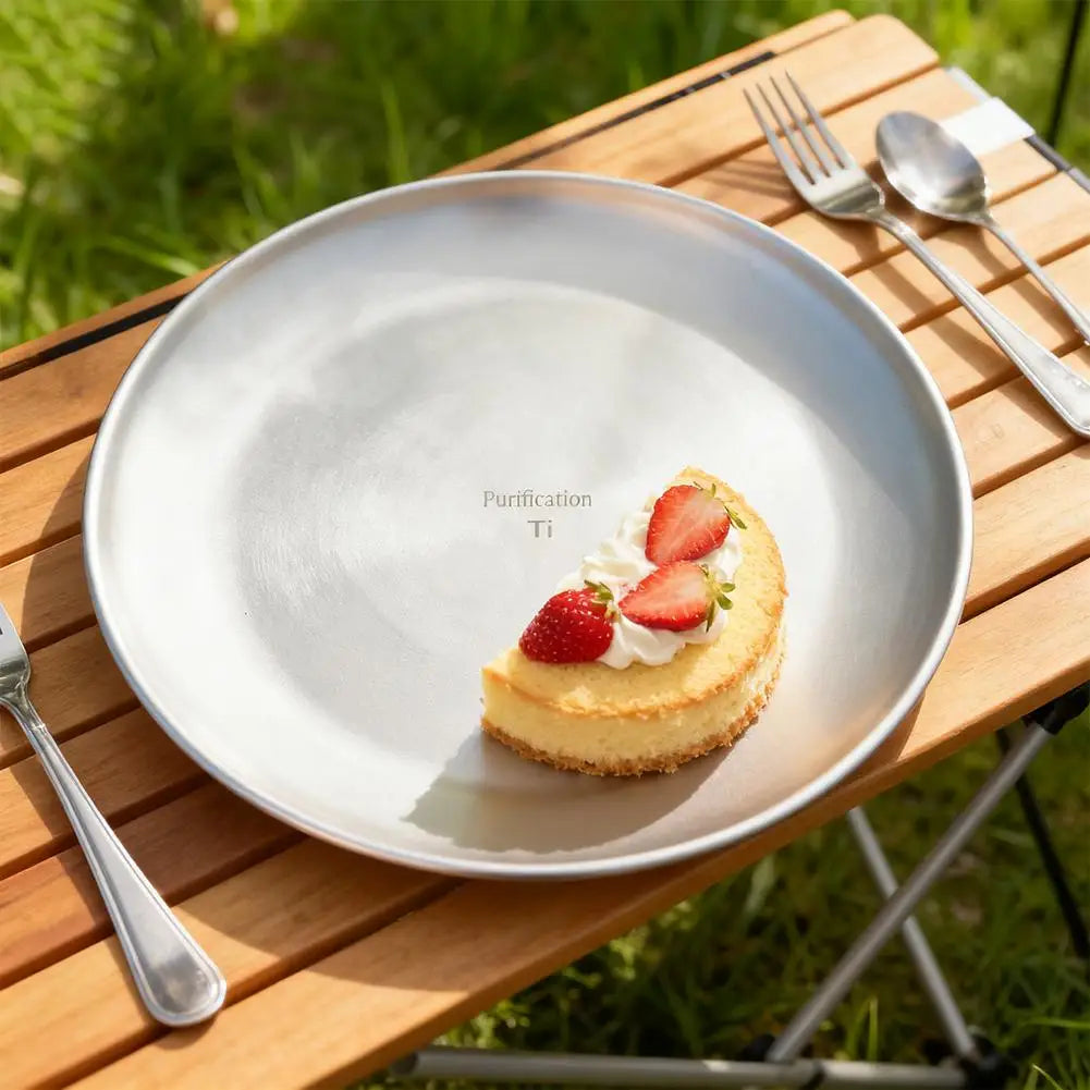 A silver titanium plate holds a cheesecake slice with whipped cream and strawberries on a wooden table outdoors.