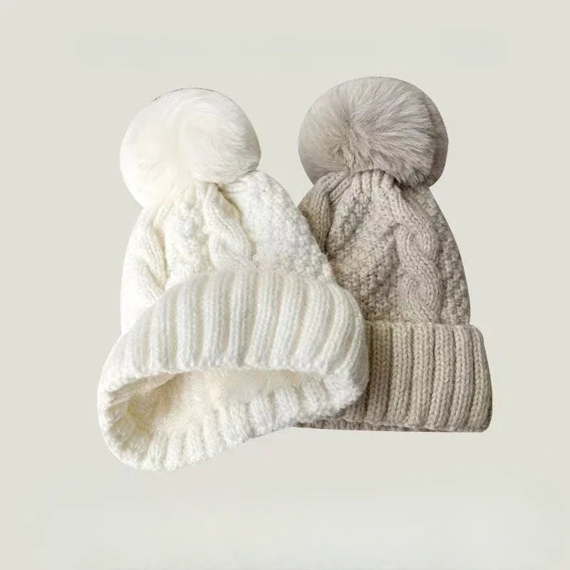Two cable-knit winter beanie hats with faux fur pom-poms, one in white and one in beige.