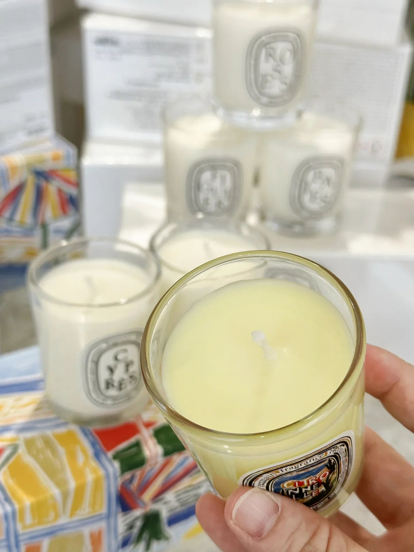 A hand holds a small glass jar candle with pale yellow wax, with other similar candles blurred in the background.