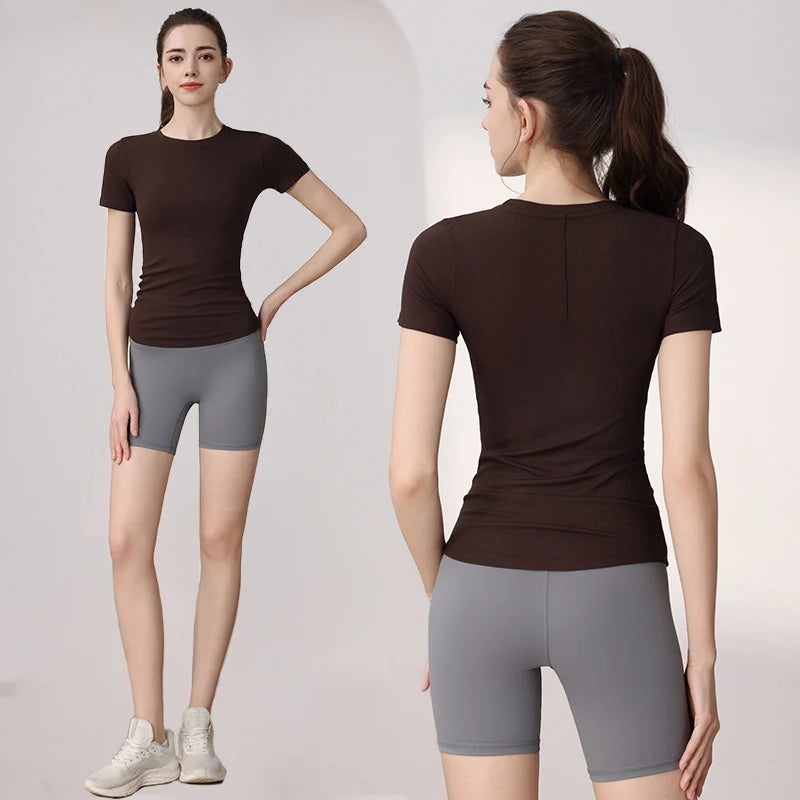 A woman wearing a dark brown short-sleeved fitted athletic top and high-waisted grey sports shorts.