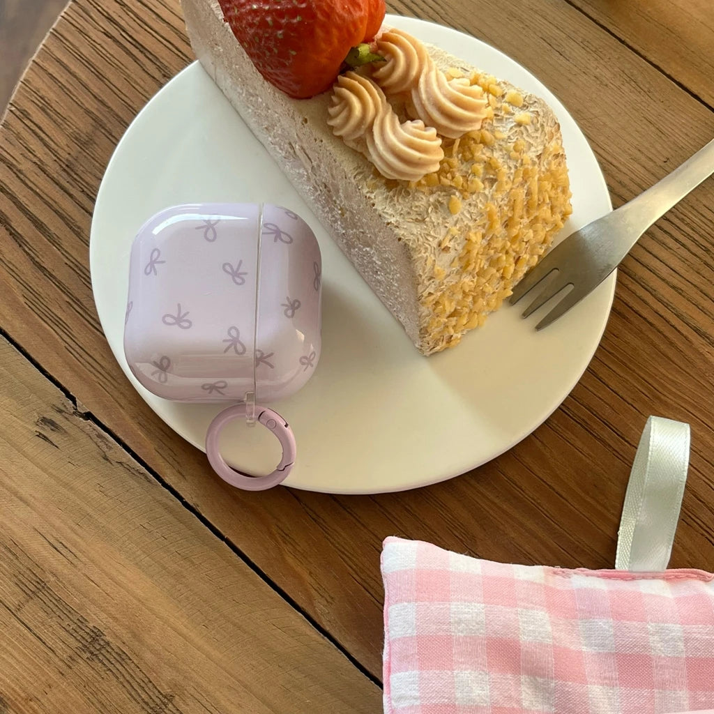 Lilac AirPods case with bow pattern and matching ring clip, next to a slice of cake on a plate.
