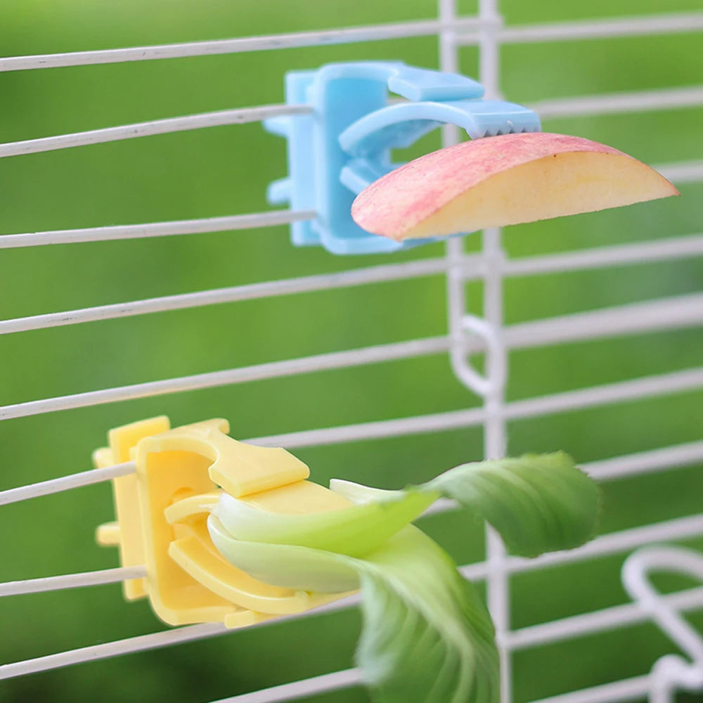 Blue and yellow plastic food clips attached to a white wire cage, holding a slice of apple and a leafy vegetable.