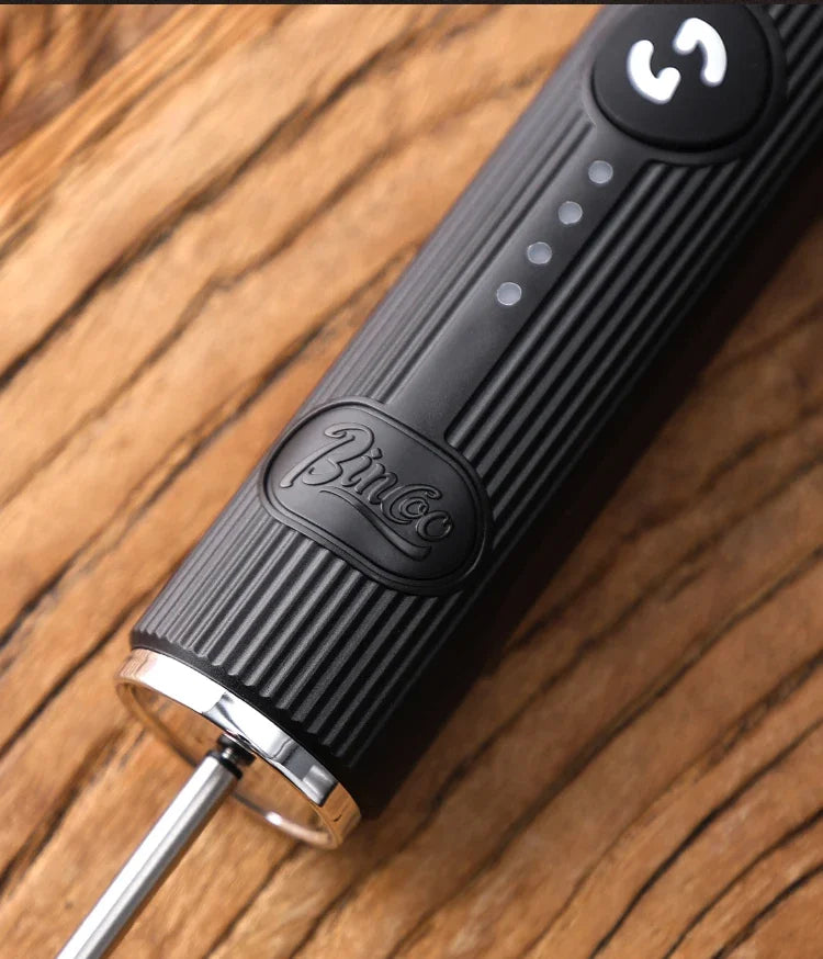 Matte black electric handheld milk frother with ribbed grip, speed controls, and a metal whisk on a wooden surface.