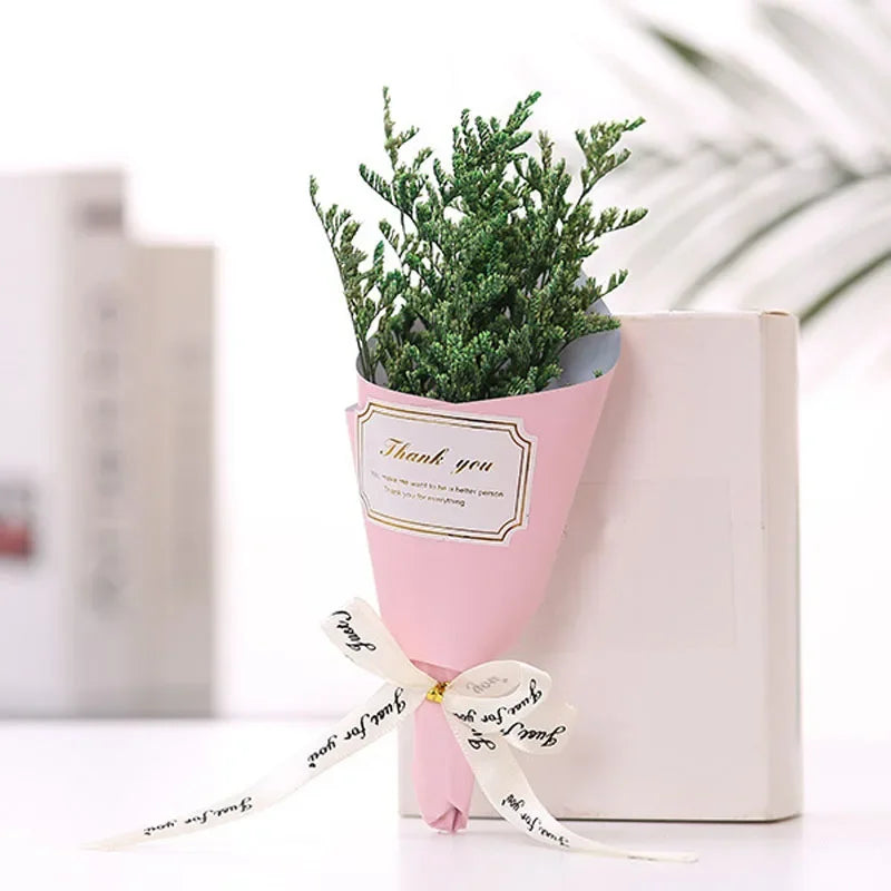 A small bouquet of artificial green sprigs wrapped in pink paper with a 'Thank you' label and a decorative ribbon.