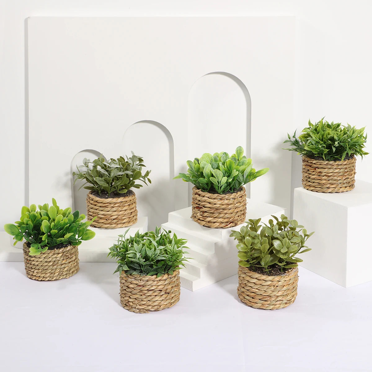 Six small artificial green plants in woven rope pots arranged on white tiered pedestals against an arched background.