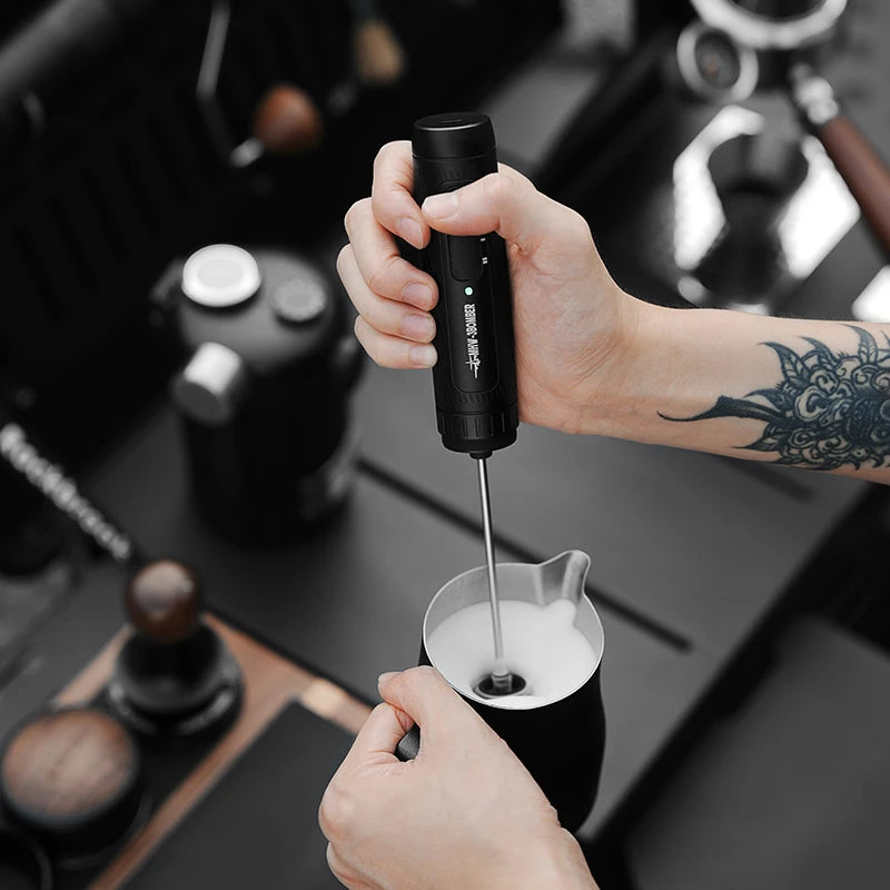 A hand holding a sleek, black electric milk frother, mixing foam in a stainless steel pitcher.
